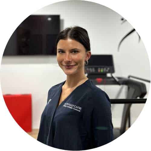 Female healthcare professional in navy scrubs smiling in a clinic with gym equipment in the background.